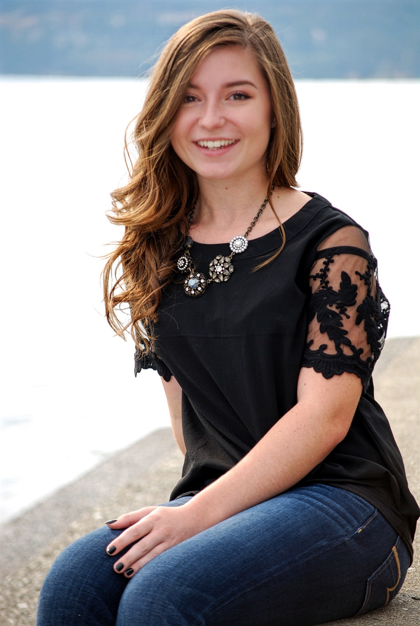 Lauren Brownlee is a young woman with long, wavy brown hair and light skin, smiling while sitting outdoors near a body of water. She is wearing a black blouse with lace details on the sleeves, a statement necklace with decorative gems, and dark blue jeans. The blurred background features a calm lake and distant mountains, suggesting a serene and scenic location.