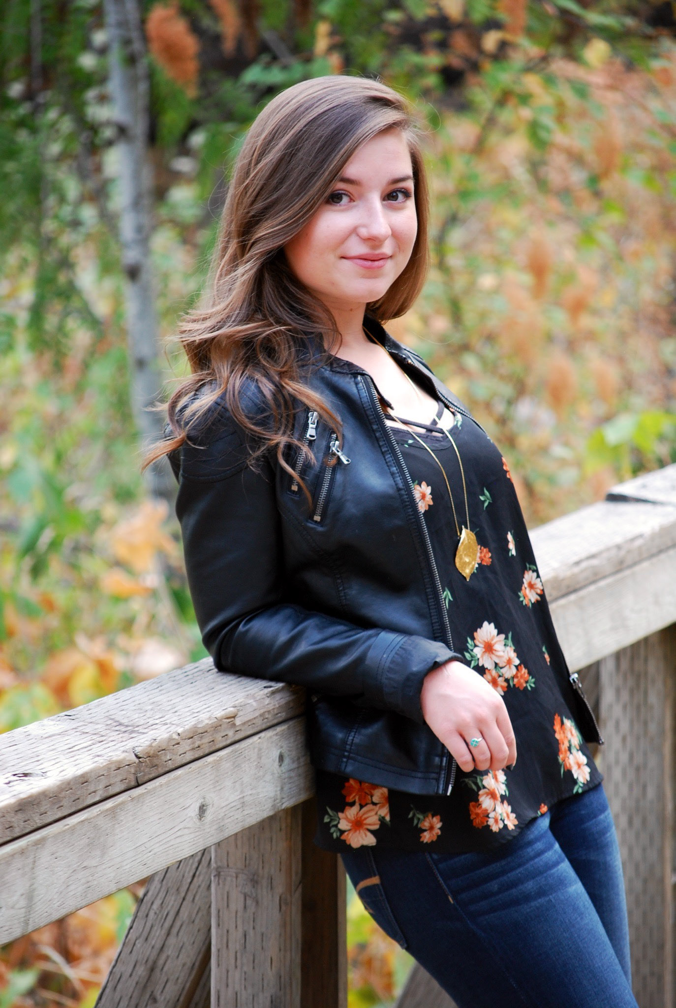 Lauren Brownless, a young woman with long, wavy brown hair and light skin, leaning against a wooden railing on a bridge in an outdoor setting with lush green and autumn-colored foliage. She is wearing a black leather jacket over a floral-patterned blouse, blue jeans, and a gold pendant necklace. She has a subtle smile and a confident expression, looking at the camera with a relaxed posture.