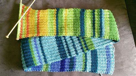 A colorful, hand-knitted scarf featuring a striped pattern in shades of green, blue, yellow, and orange, displayed on a soft brown surface. The knitting needles, still attached to the scarf, rest on the fabric, indicating that the project is in progress. The texture of the scarf is ribbed, with alternating bands of knit and purl stitches, creating a warm and stretchy fabric. The vibrant colors transition smoothly, suggesting the use of variegated yarn.