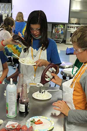 Two campers wearing aprons and putting chocolate sauce on whipped cream in an educational kitchen