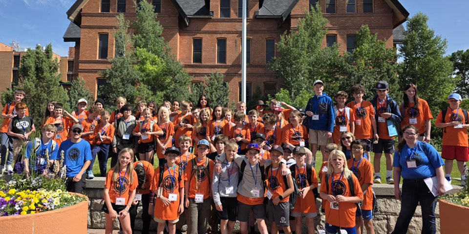 A group of smiling children and camp staff pose together outside a sunny brick building on MSU's campus. They are wearing name tags and their Peaks tee-shirts.